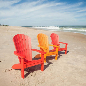 Three child size Adirondack chairs in red, orange and pink lined up on the sand facing breaking ocean waves