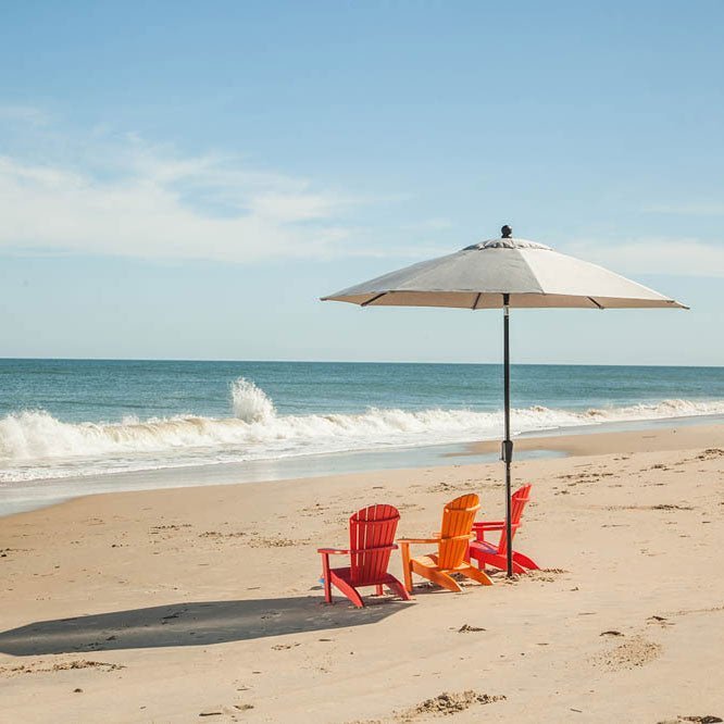 Red, orange and pink child size Adirondack chairs grouped under a market umbrella on an empty beach