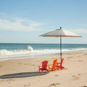 Red, orange and pink child size Adirondack chairs grouped under a market umbrella on an empty beach