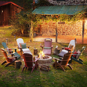 Ring of colorful Adirondack chairs with pillows around a blazing backyard fire pit beside a stone farmhouse