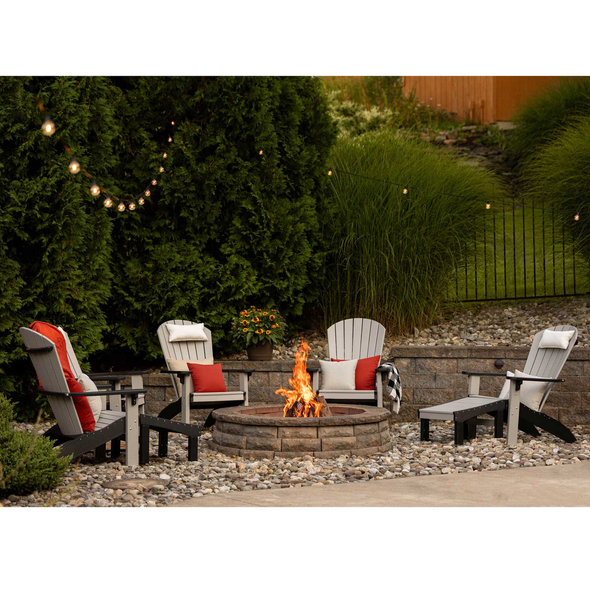 Gray and black Adirondack chairs with red and white pillows and an ottoman circling a lit stone fire pit at dusk