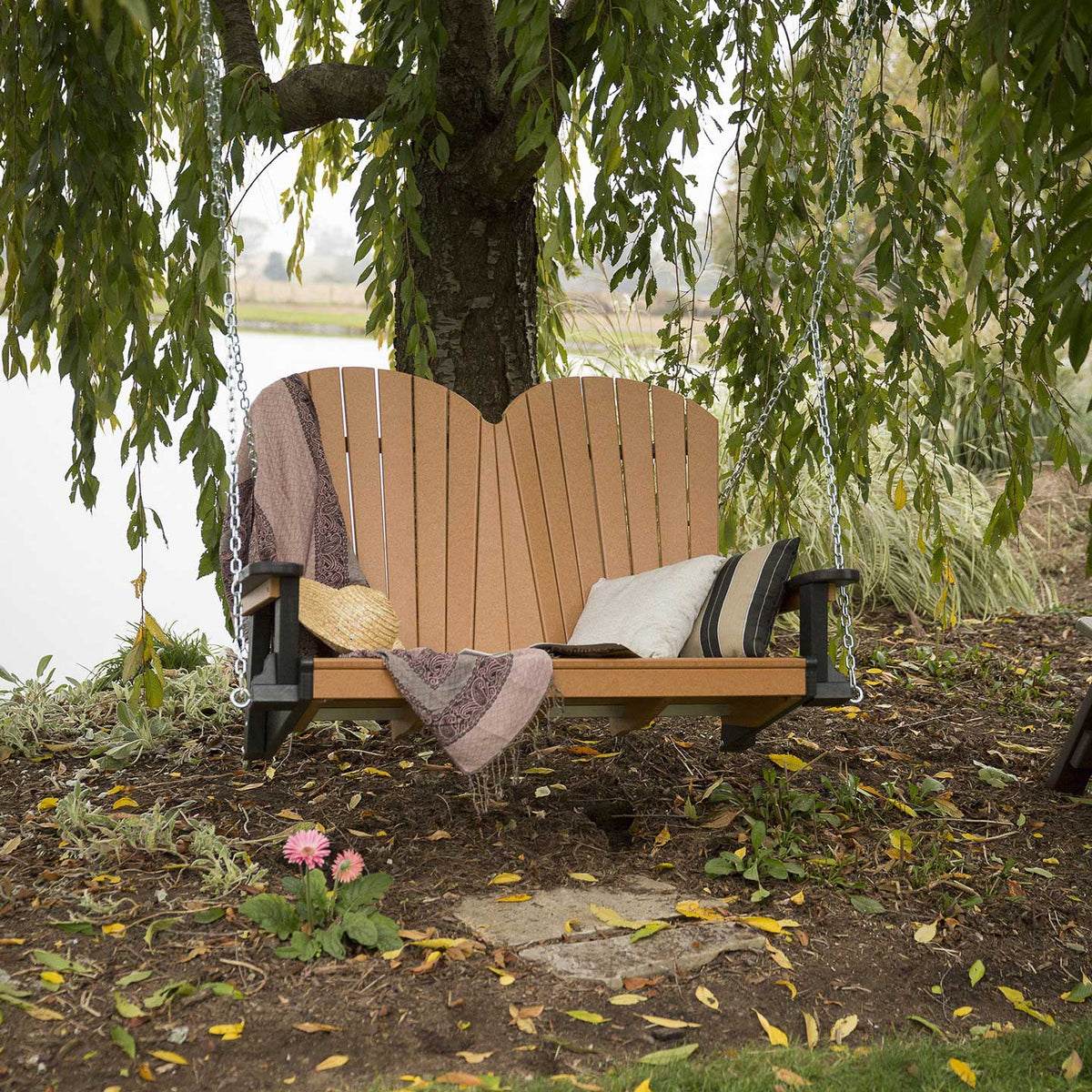 Cedar and black poly porch swing hung from a willow tree by a pond, styled with pillows, a quilt and straw hat