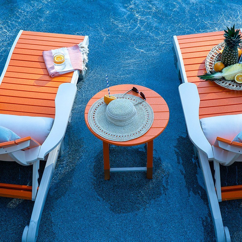 Overhead view of an orange round poly side table with a sun hat and drink between two chaise lounges by a pool