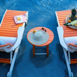 Overhead view of an orange round poly side table with a sun hat and drink between two chaise lounges by a pool
