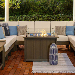 Driftwood gray square propane fire pit table burning between white cushioned sectional seating on a brick patio