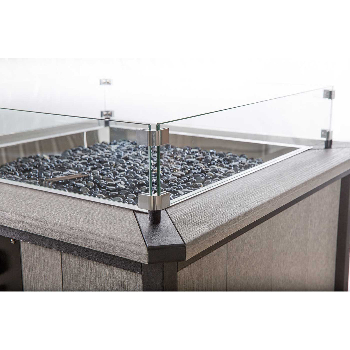 Corner detail of a gray poly fire pit table showing the stainless clip holding the glass guard above black lava stones