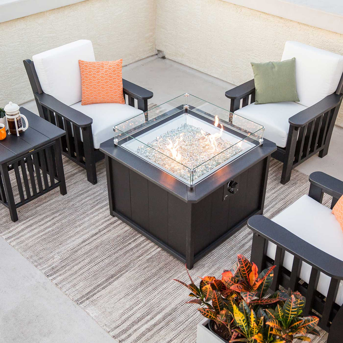 Black square fire pit table with clear glass beads and flames ringed by black mission chairs with white cushions