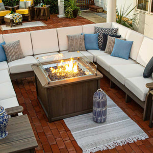 Gray poly square fire pit table with tall flames and glass guard anchoring a large L-shaped outdoor sectional
