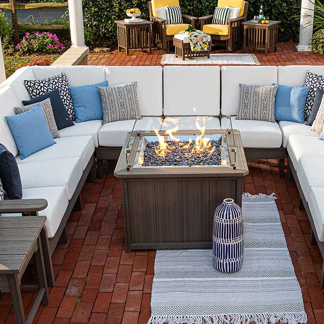 Overhead view of a gray square fire pit table with dark fire glass surrounded by a white cushioned sectional on brick