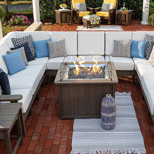 Overhead view of a gray square fire pit table with dark fire glass surrounded by a white cushioned sectional on brick