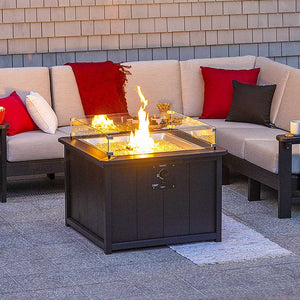Square black poly propane fire pit table with a glass wind guard burning beside a cream cushioned sectional