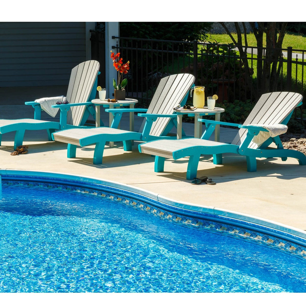 Three driftwood gray and blue poly sun lounge chairs with ottomans and side tables lined along a pool deck
