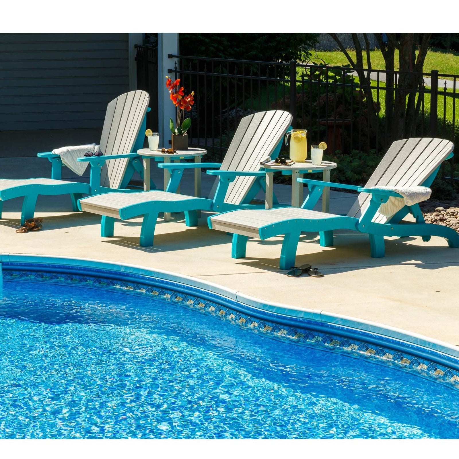 Row of driftwood and aruba blue poly chaise lounges with round side tables reflected in a sunny swimming pool
