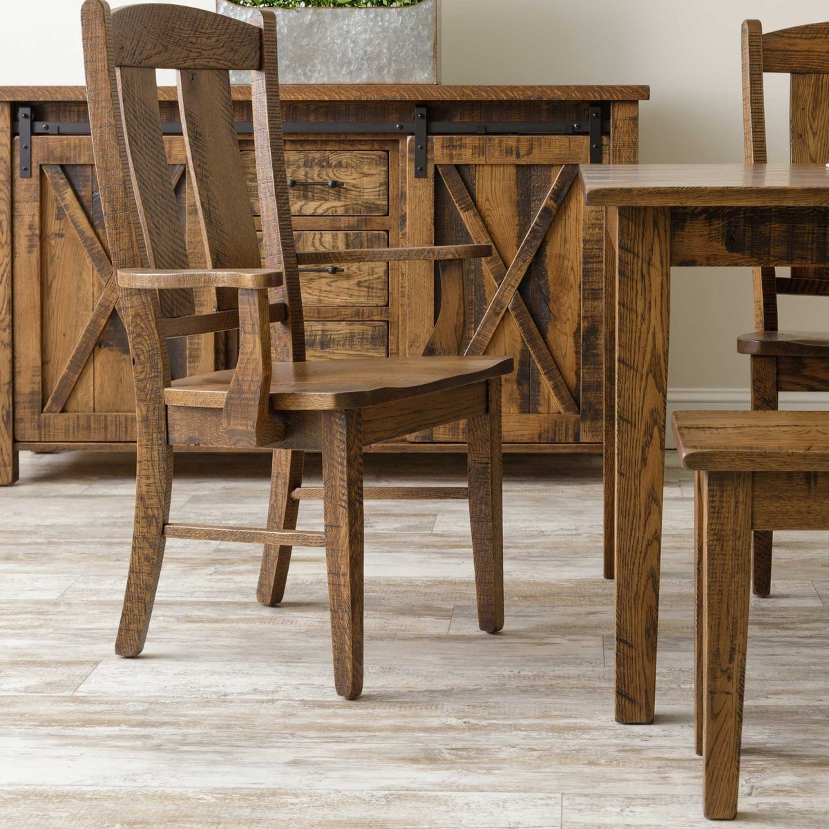 Rustic oak dining chair with arms next to a barn door sideboard, showing the thick square table leg and scooped wood seat