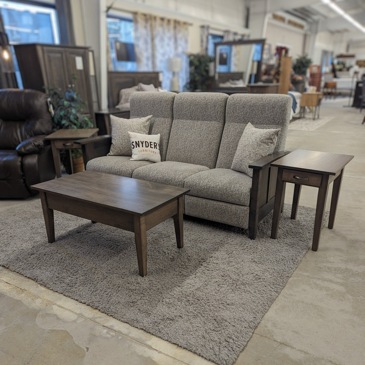 Gray tweed reclining sofa on a showroom floor with a Snyder's Furniture pillow, dark coffee table and end table
