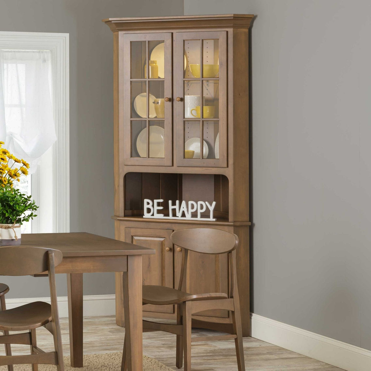 Lighted corner china cabinet in a light brown finish with glass mullion doors, open shelf and lower storage doors in a dining nook