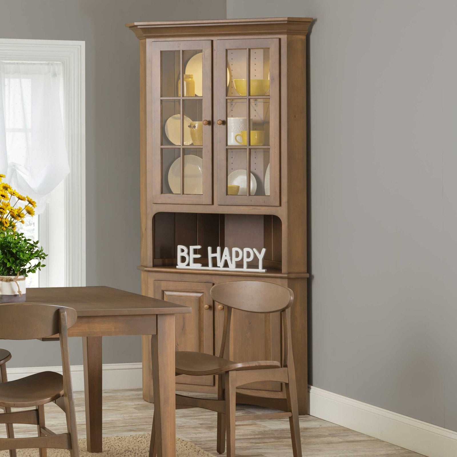Lighted corner china cabinet in a light brown finish with glass mullion doors, open shelf and lower storage doors in a dining nook