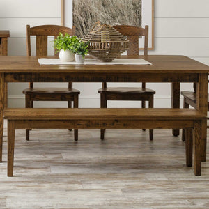 Rustic oak dining bench with a plank seat and tapered legs, tucked under a matching farmhouse table with greenery and a basket