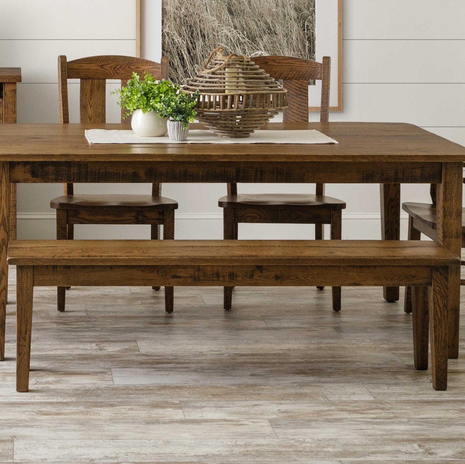 Rustic oak dining bench with a plank seat and tapered legs, tucked under a matching farmhouse table with greenery and a basket