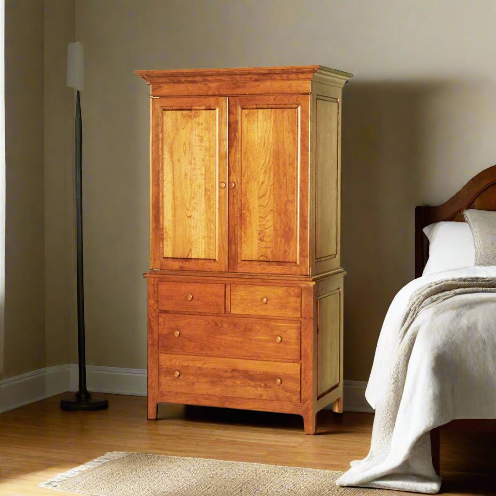 Tall cherry armoire with two raised panel doors over four drawers and crown molding beside a bed