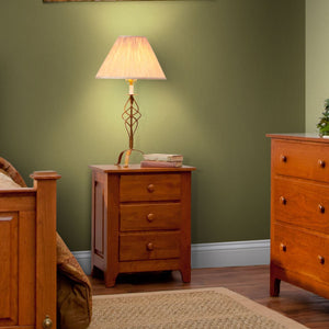 Three drawer cherry nightstand with round knobs under a lamp in a green painted bedroom
