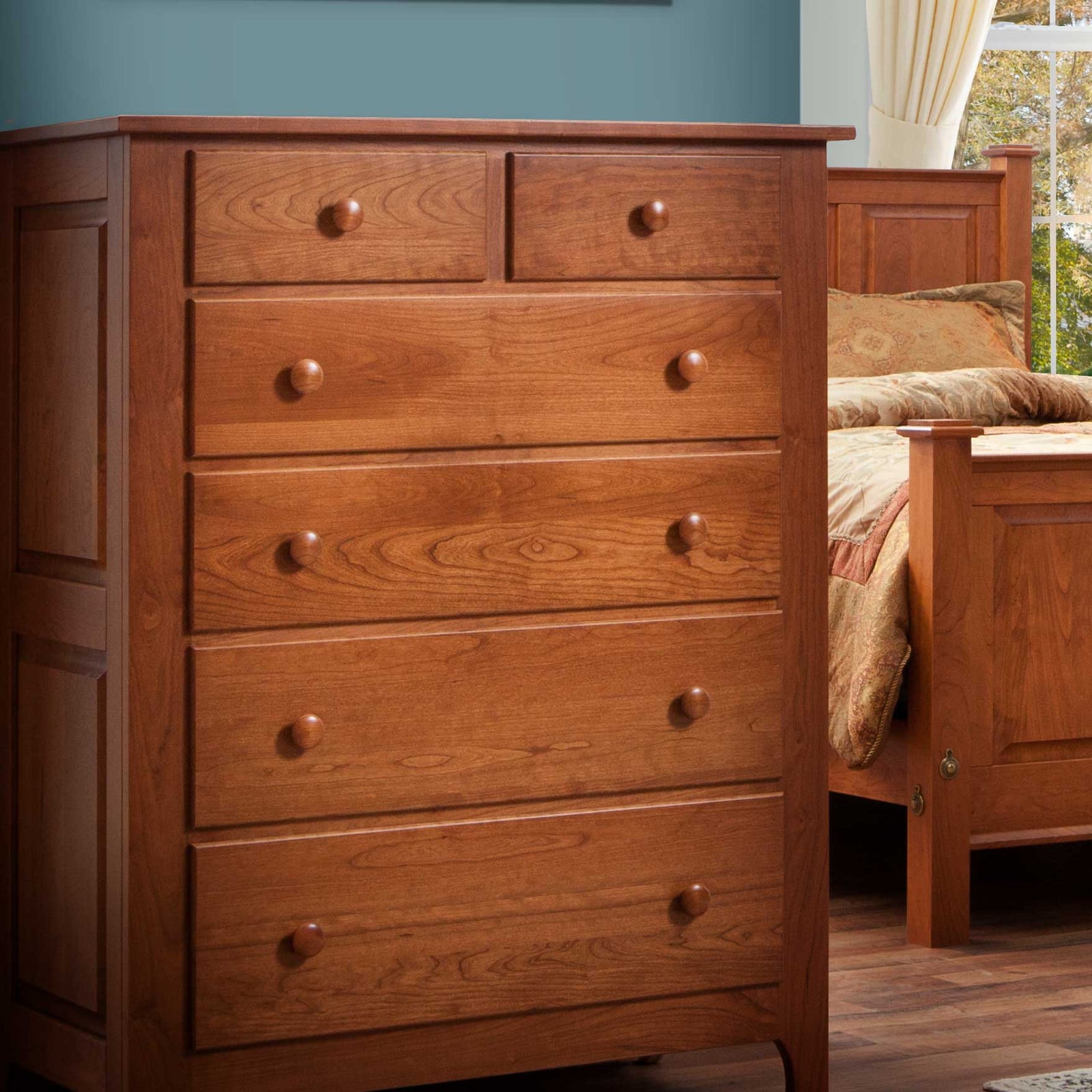 Cherry chest of six drawers photographed in a bedroom beside a panel bed with patterned bedding
