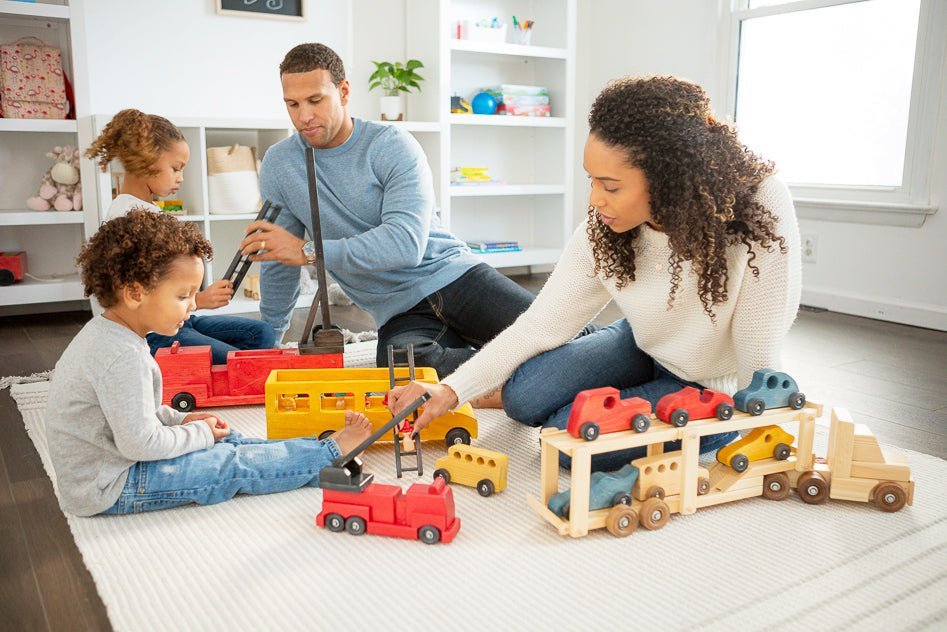 Family sitting on a rug playing with an assortment of wooden toy trucks, cars and a red fire engine