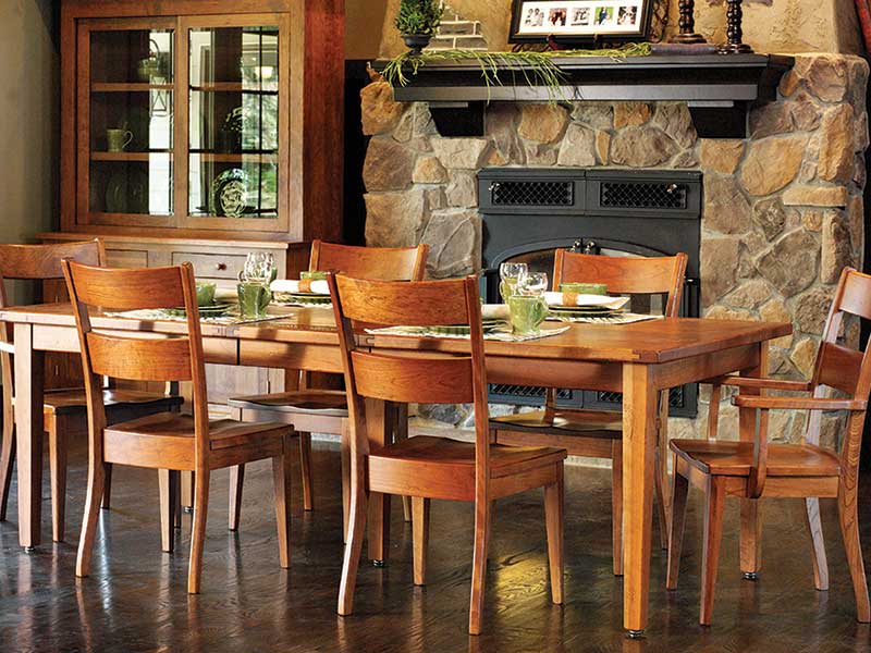 Cherry dining table with wood seat side and arm chairs set with glassware near a stone hearth