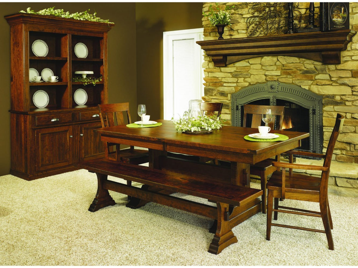 Rustic cherry trestle dining table with bench, slat back chairs and a hutch by a stone fireplace