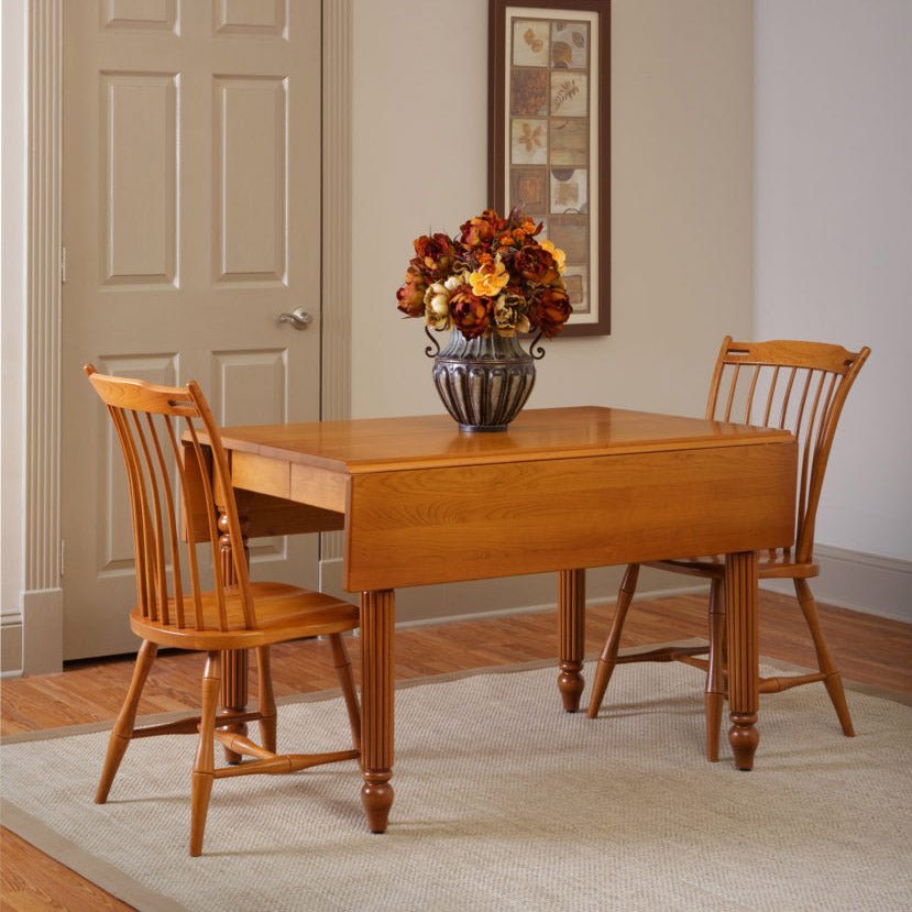 Oak drop leaf dining table with one leaf down and two spindle back chairs beside a floral arrangement in a hallway