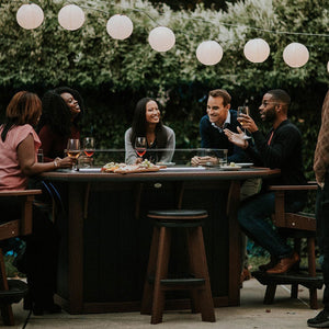 Group of friends seated on stools at a dark brown poly fire pit bar table with wine under hanging lanterns