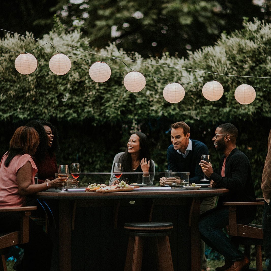 Friends gathered with wine around a brown poly fire pit counter table under strung paper lanterns