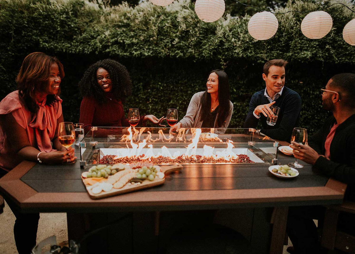 Five friends laughing around a lit linear fire trough on a brown poly bar table with a cheese board