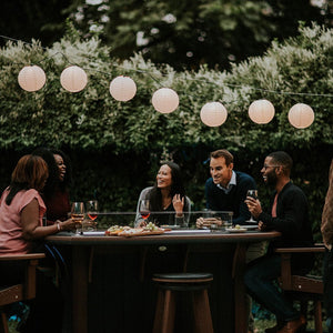 Backyard party of five seated at a dark brown poly fire pit table with wine under paper lanterns