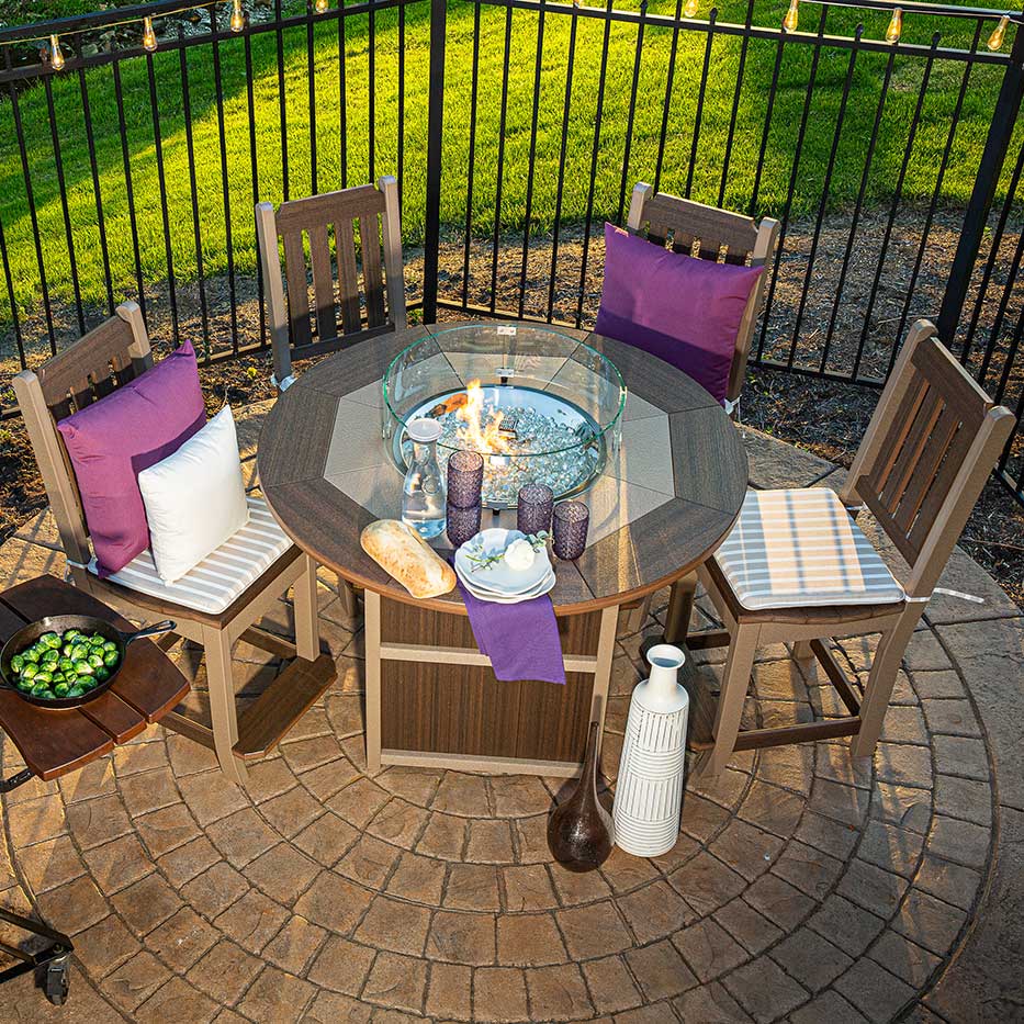 Overhead view of a round poly fire pit counter table with lit flames, four slat back bar chairs and purple accent pillows on a stone patio