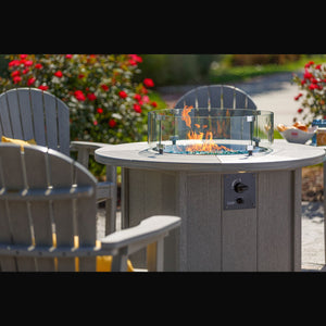 Lit gray poly fire pit table with turquoise fire glass and control knob, surrounded by fan back Adirondack chairs on a sunny patio