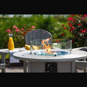 Driftwood gray poly round fire table with a glass wind guard over blue fire glass, flanked by gray Adirondack chairs and rose bushes