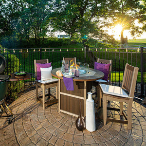 Brown and tan poly counter height fire pit table with a glass flame guard, set with four cushioned bar chairs on a stamped patio at sunset