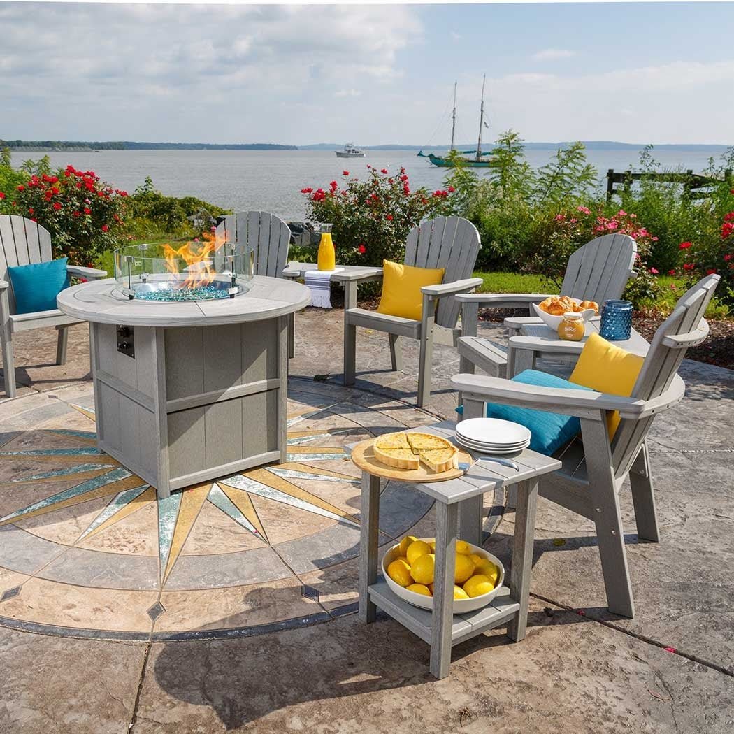 Bayside patio set with a gray poly round fire pit dining table, six Adirondack chairs, side tables and a sailboat view