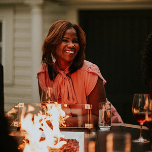 Guests gathered at a lit outdoor fire table, one smiling woman in coral with wine glasses behind the glass wind guard