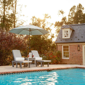 Two gray poly sun loungers with white cushions and a market umbrella beside a pool by a brick pool house at sunset