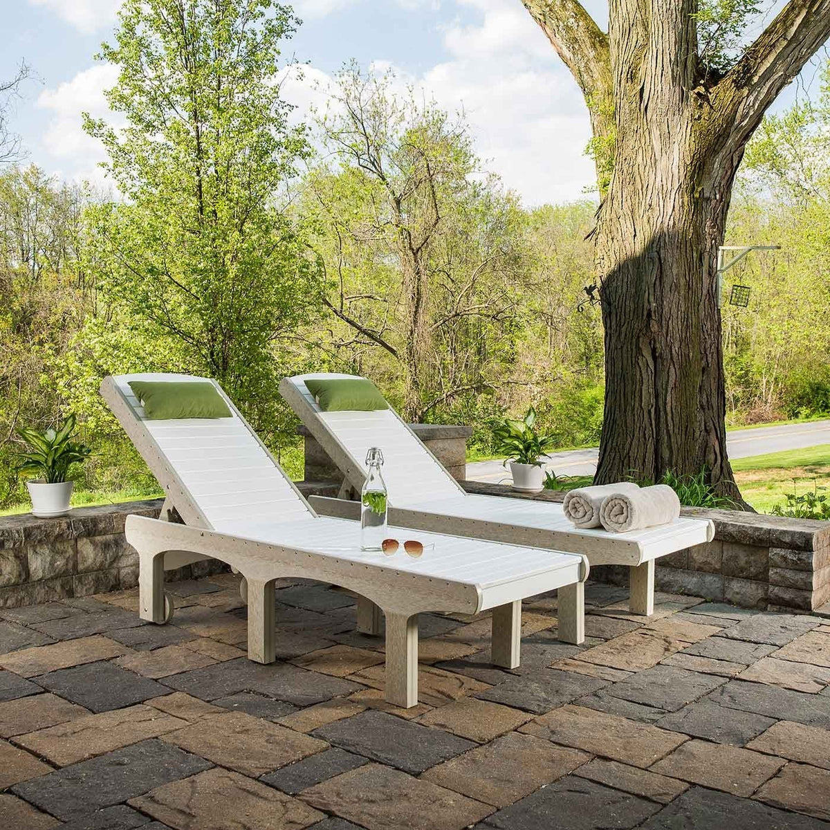 Two white poly sun loungers with green pillows, a water bottle and rolled towels on a paver patio under a shade tree