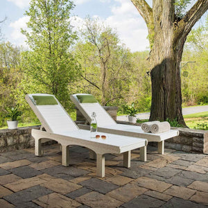 Two white poly sun loungers with green pillows, a water bottle and rolled towels on a paver patio under a shade tree