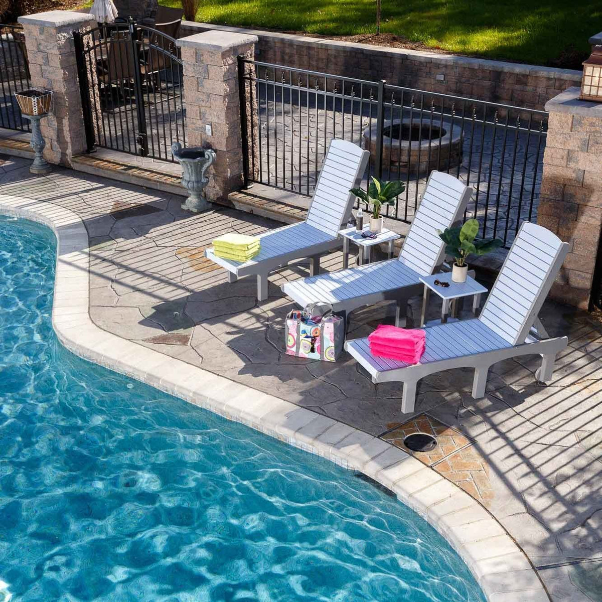 Poolside view of three gray poly chaise lounges with side tables, bright towels and a beach bag on a flagstone deck