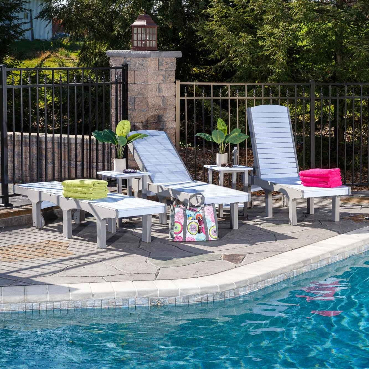 Gray and white poly sun loungers with reclined and upright backs, side tables and towels beside a pool and iron fence