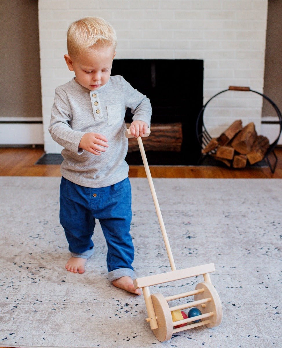 Blond toddler in a gray shirt pushing a wooden block roller push toy across a rug by a brick fireplace