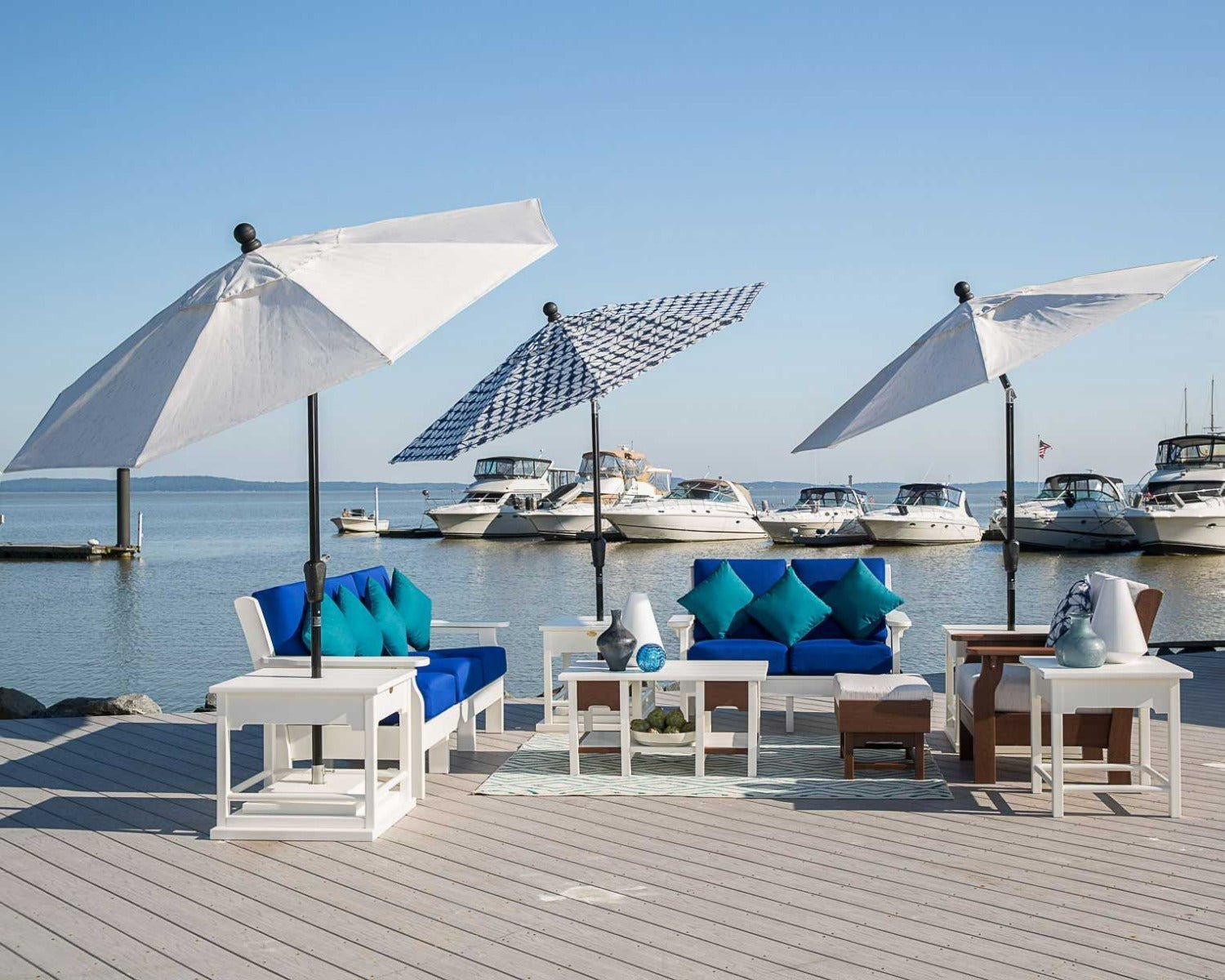 Marina dock lounge setting with white and walnut poly sofas, blue and teal cushions, side tables and market umbrellas