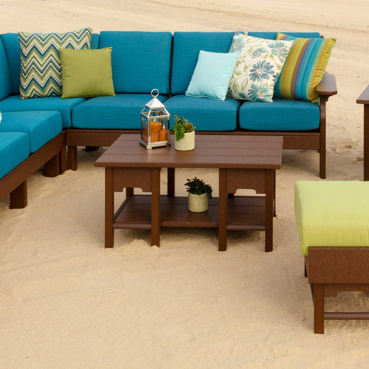 Brown poly patio coffee table with a lantern and plants, staged in sand beside a teal cushioned sectional sofa