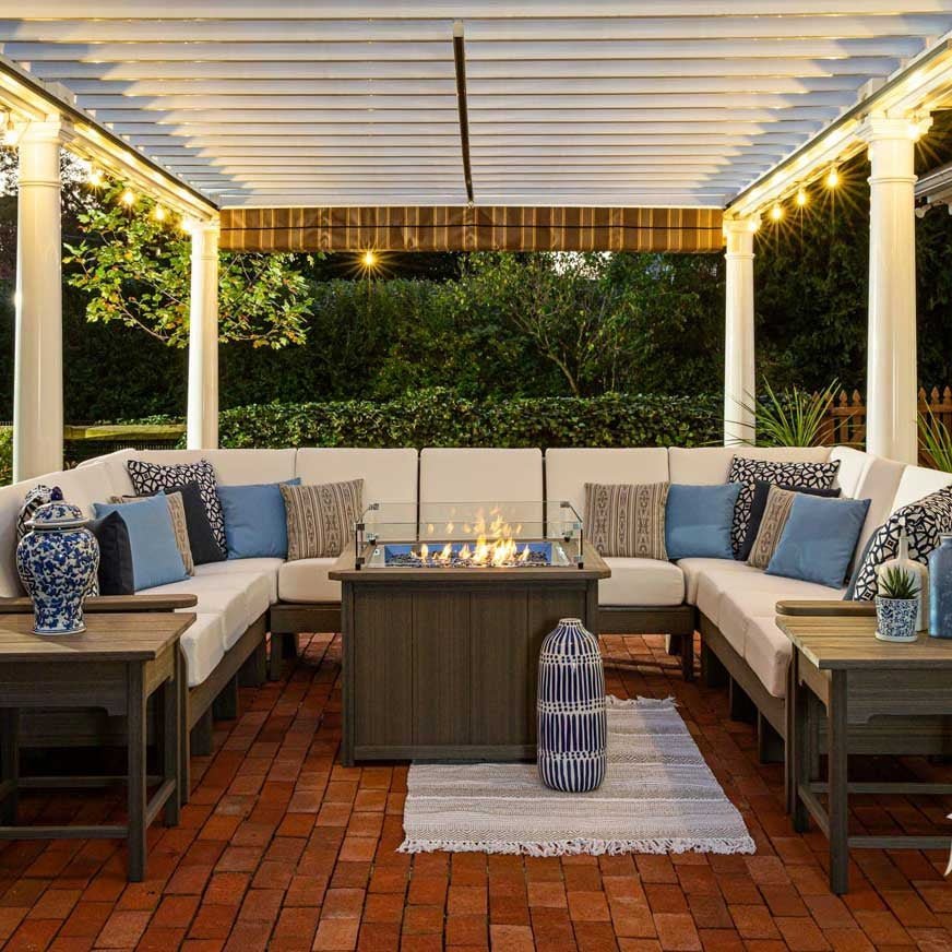 Large U shaped patio sectional with cream cushions and blue pillows around a lit linear fire table under a pergola
