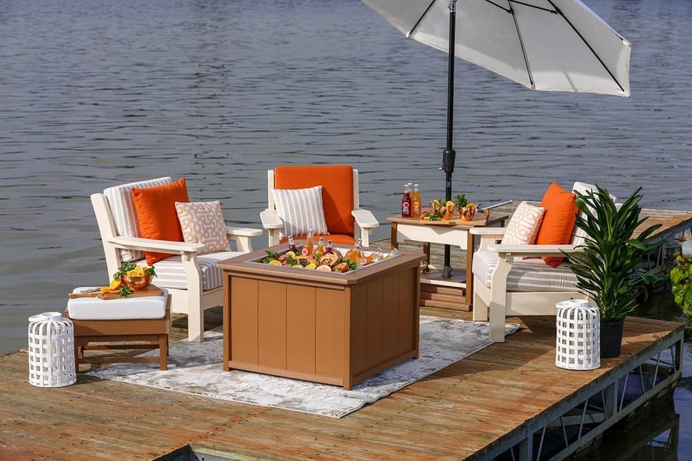 Three white poly patio chairs with orange pillows around a cedar fire pit table with food on a lakeside dock